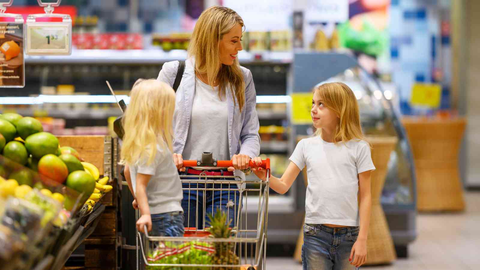 A woman with two children shops in a grocery store, pushing a cart filled with various items.
