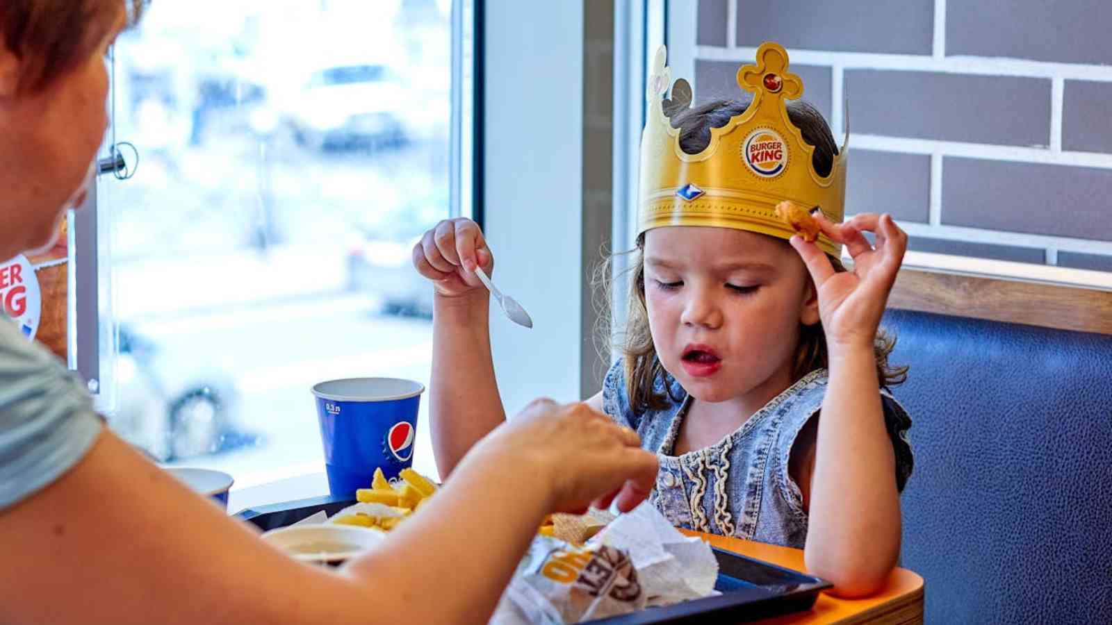 Child wearing a Burger King crown eats fries while sitting at a table with an adult. A drink cup is visible in the background.