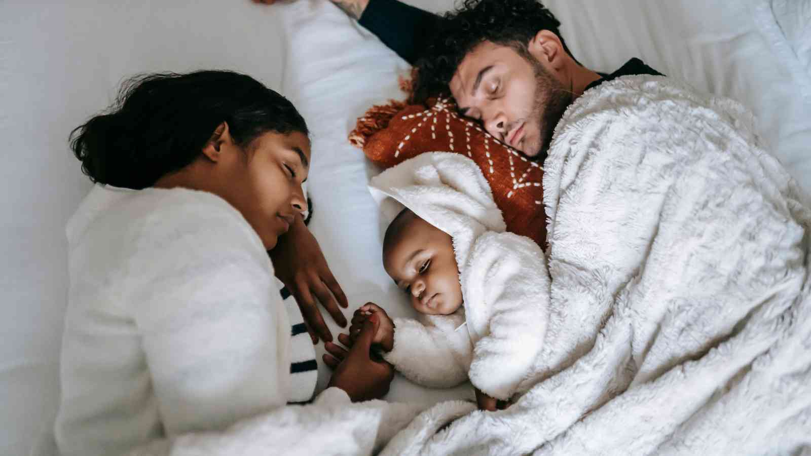 A family of three sleeps in a bed, wrapped in blankets. The child is in the middle, wearing a hooded outfit, and holding hands with the adult on the left.