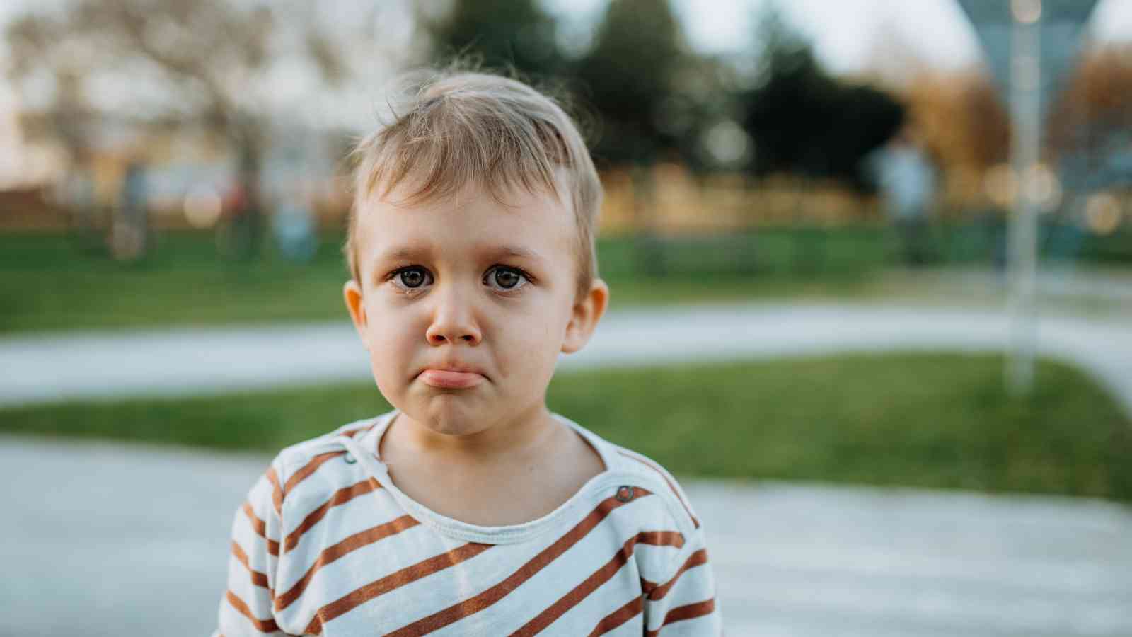 Child with a striped shirt stands outdoors with a pouty expression, blurred greenery in the background.