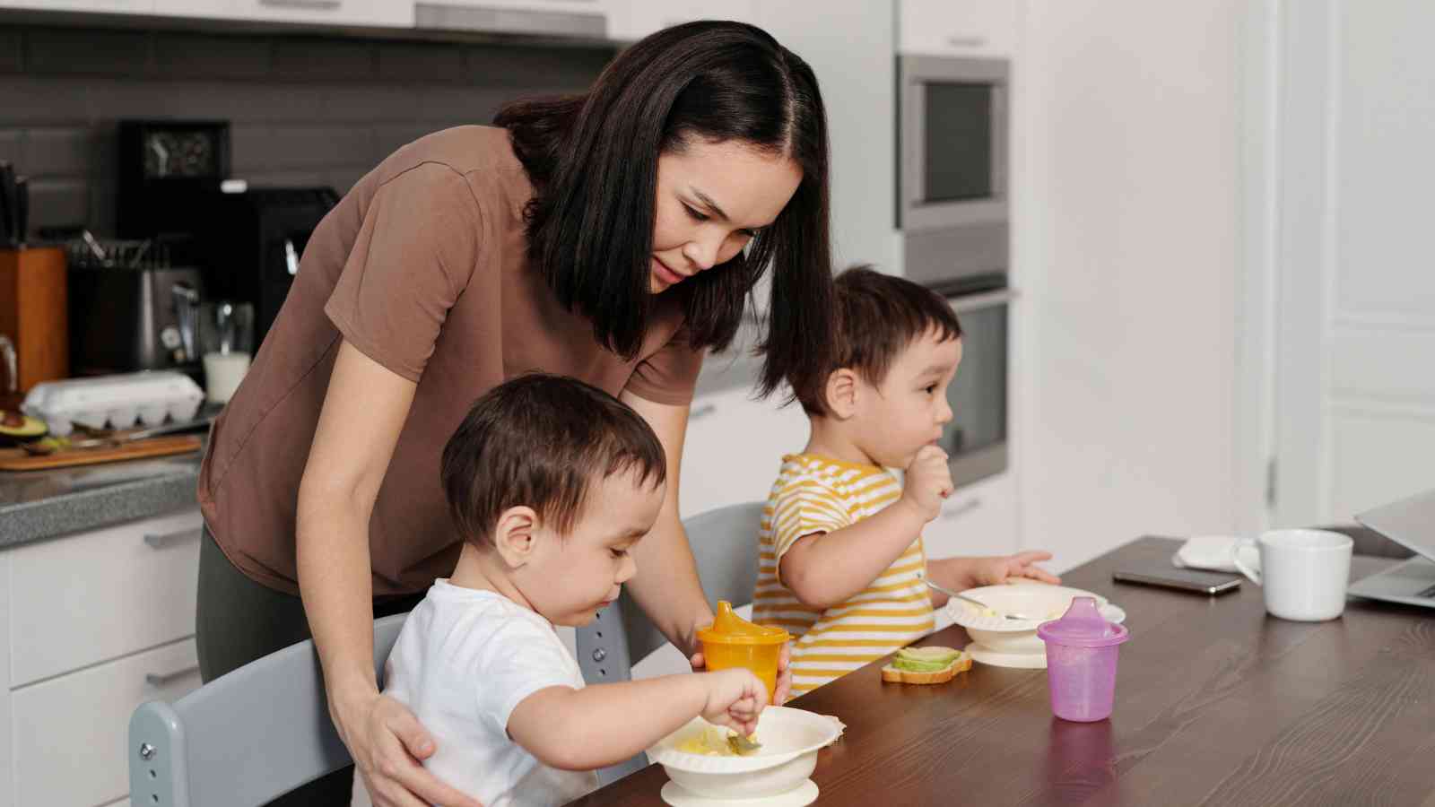 A woman helps two toddlers eat at a kitchen table. The children are using spoons and have cups nearby. The setting is a modern kitchen.