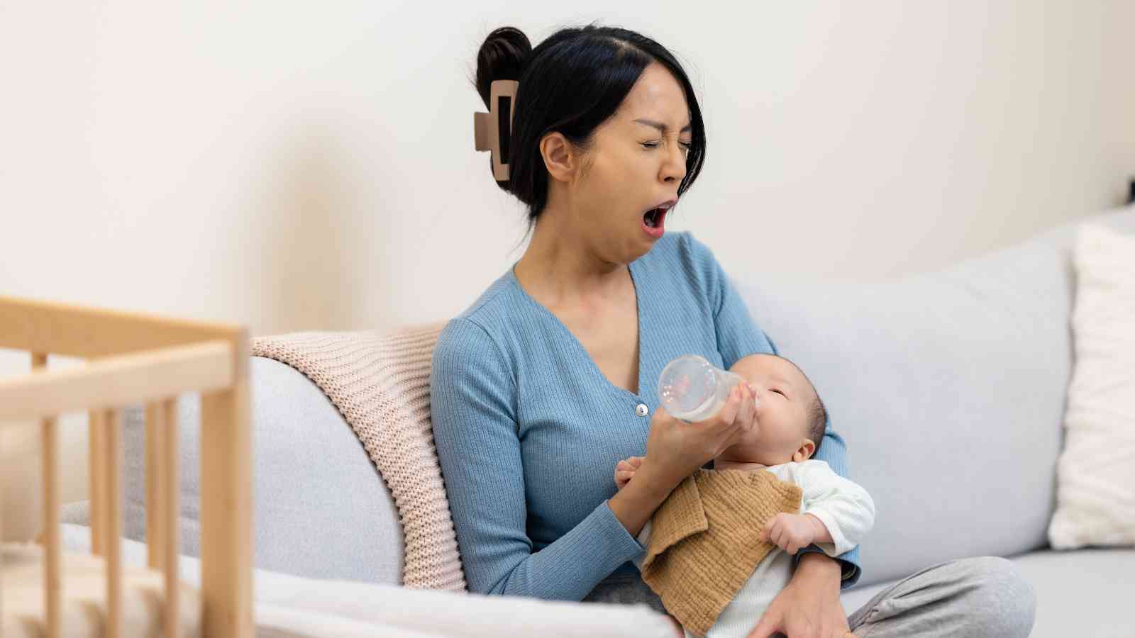 Woman yawning while feeding a baby with a bottle, sitting on a sofa next to a crib.