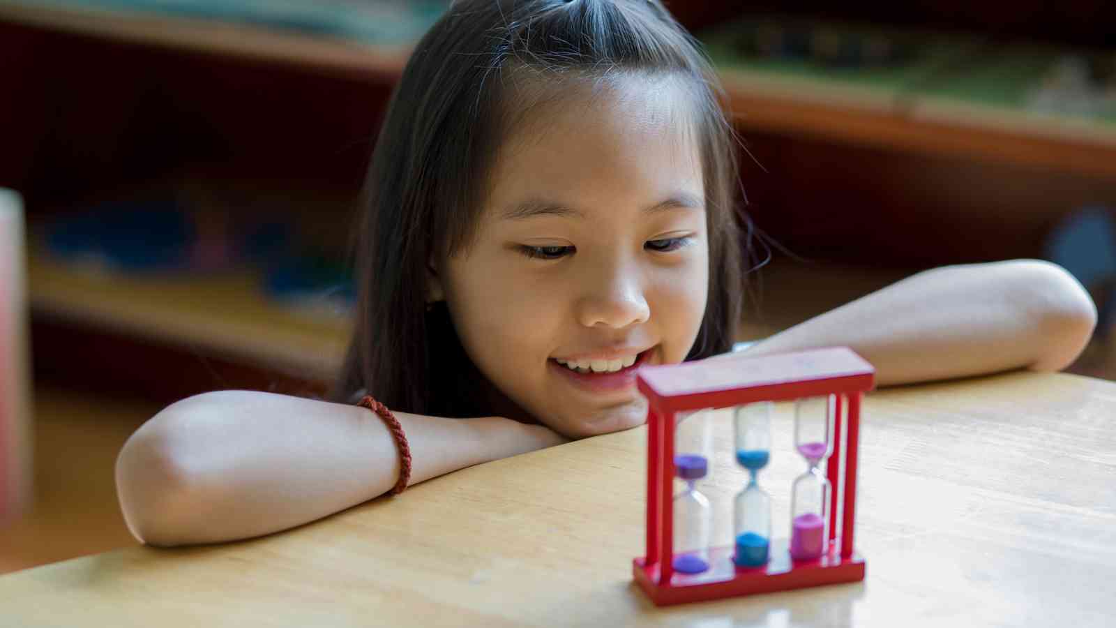 A girl smiles while watching a red-framed sand timer with blue and pink sand on a wooden table.