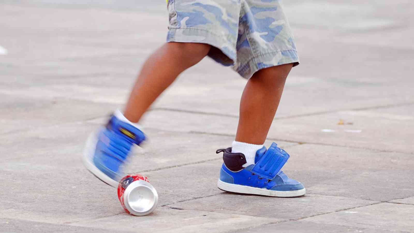 Child in camo shorts and blue sneakers kicking a crushed soda can on pavement.