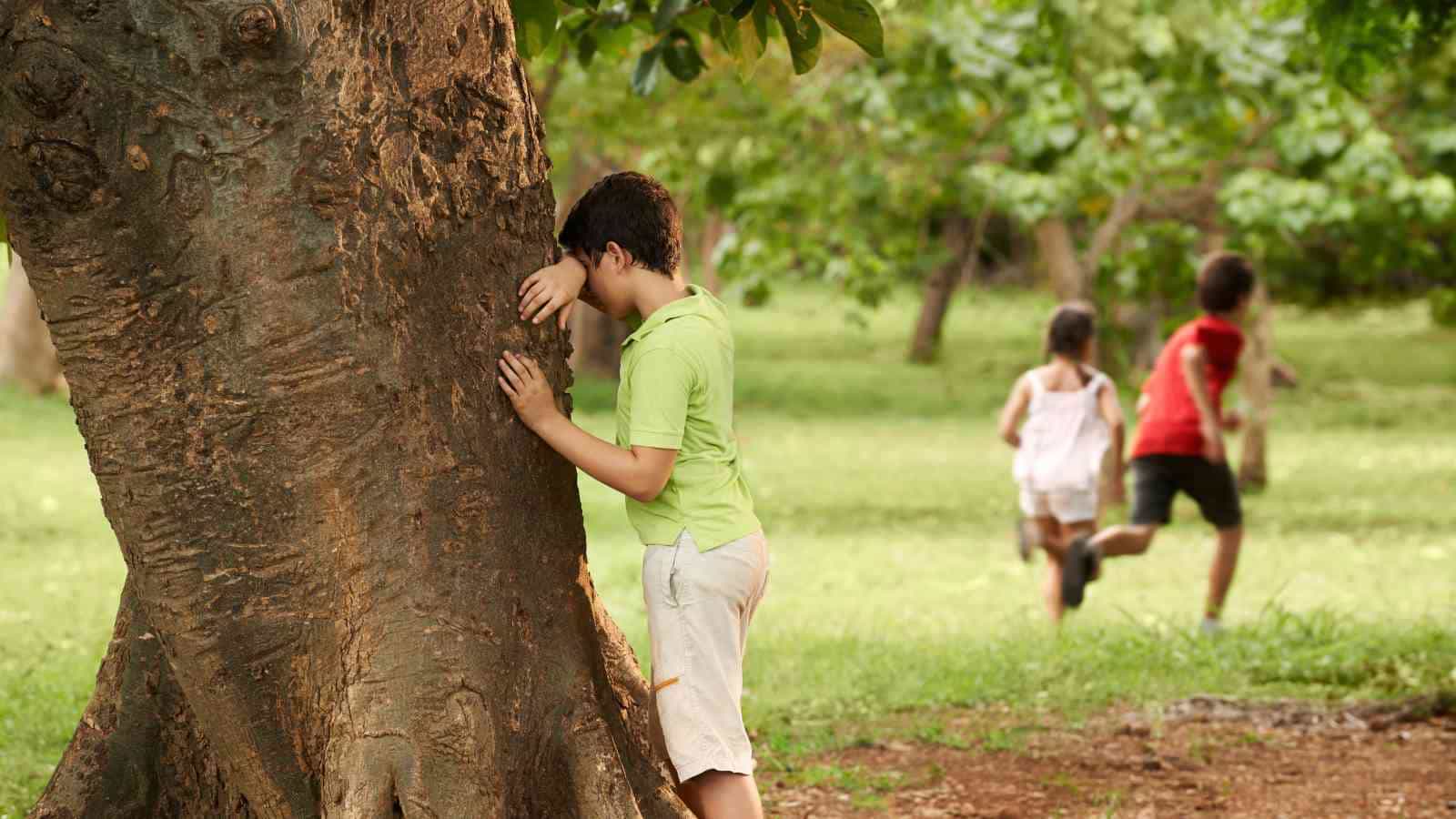 A child in a green shirt leans against a tree with eyes covered, while other children run across a grassy area in the background.