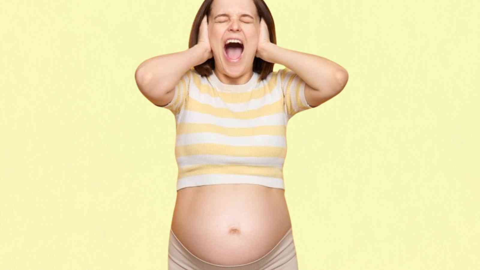 A pregnant woman screams with eyes closed and hands covering her ears, wearing a striped shirt and standing against a yellow background.