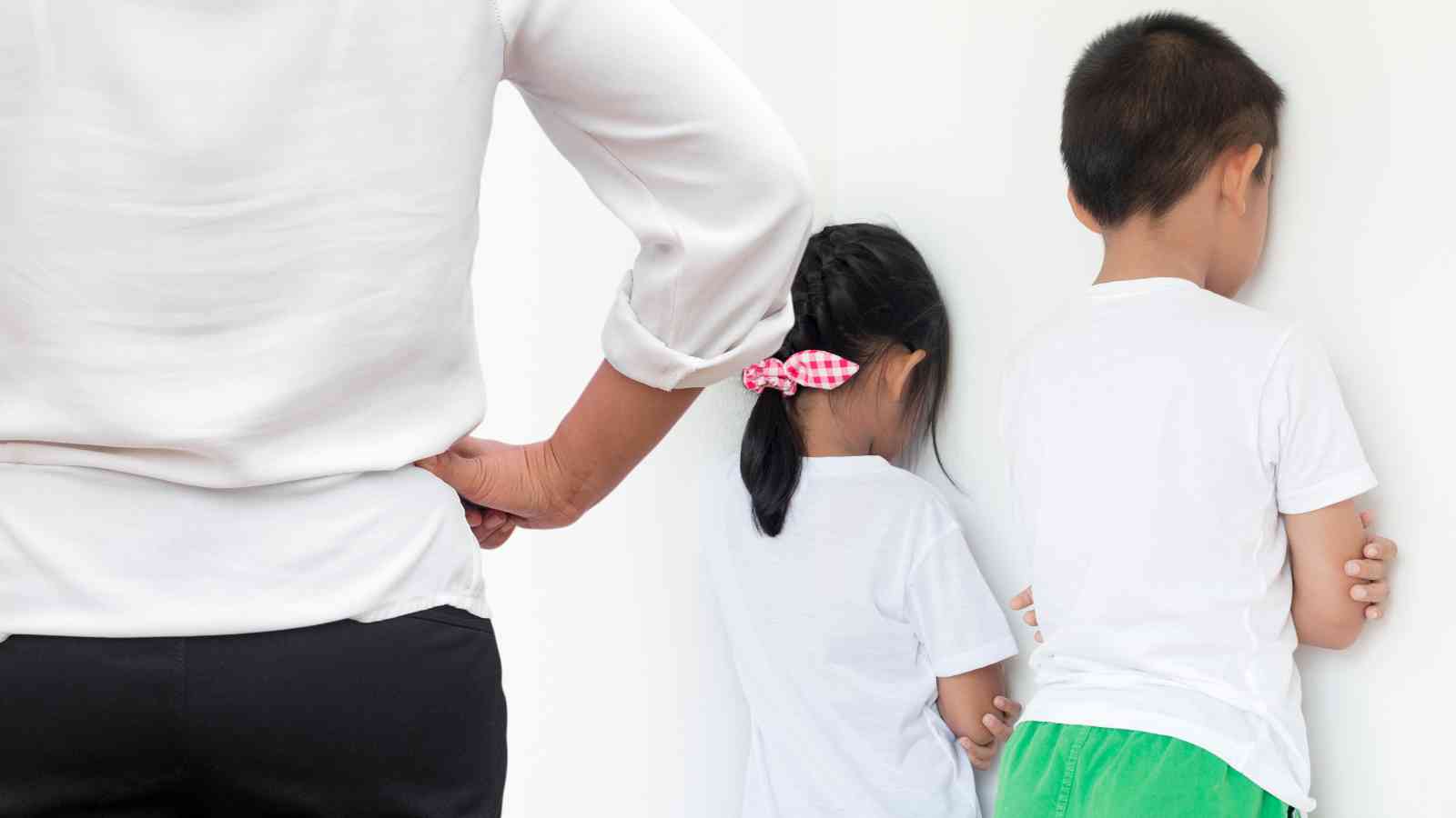Adult stands with hands on hips while two children face the wall. The girl wears a white shirt and has a pink bow; the boy wears a white shirt and green shorts.
