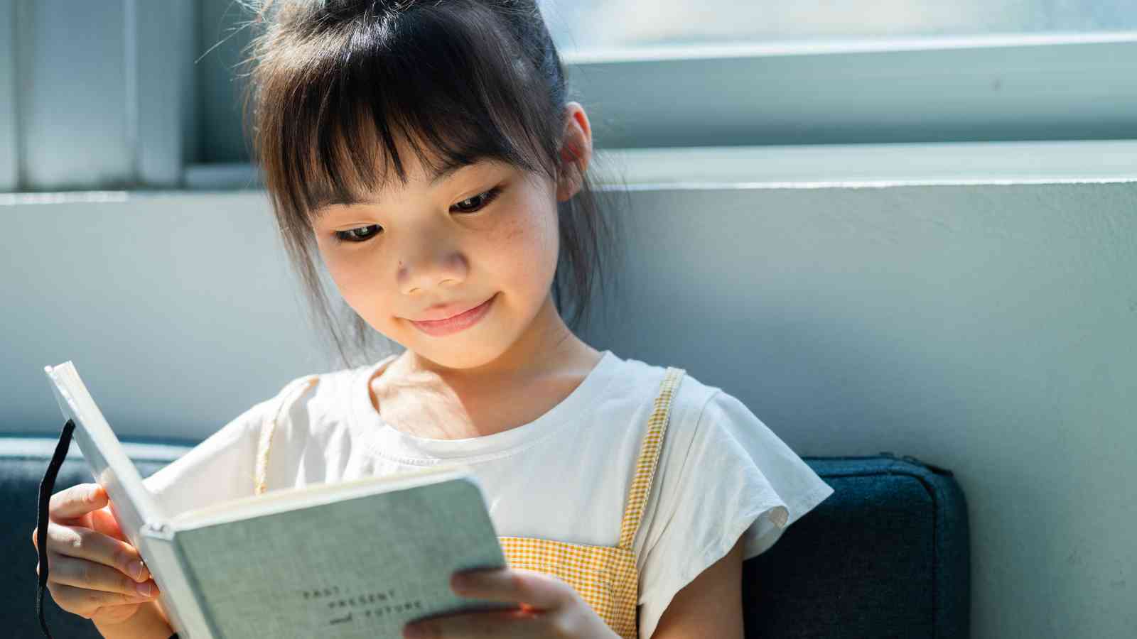 A young girl sitting by a window, reading a book with a light gray cover. She wears a white t-shirt and yellow suspenders. Daylight illuminates her face.
