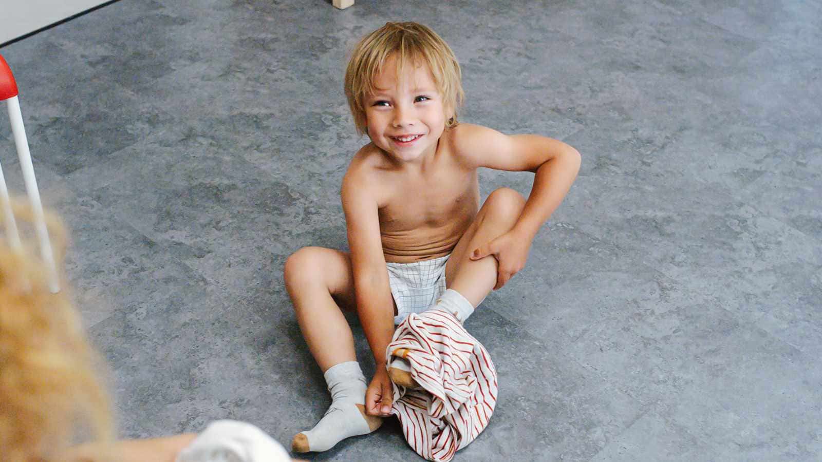 Child sitting on the floor, smiling while putting on striped pants over diaper and socks, with a red chair visible to the side.
