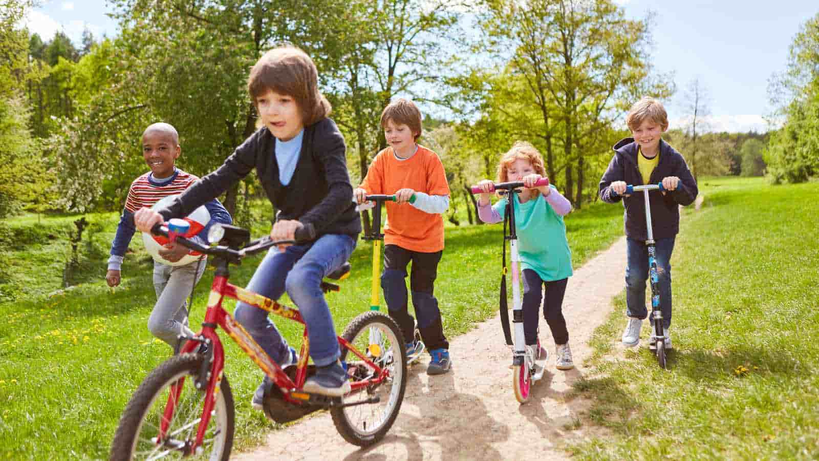 Children are riding a bicycle and two scooters on a dirt path in a park on a sunny day. Trees and grass surround them.