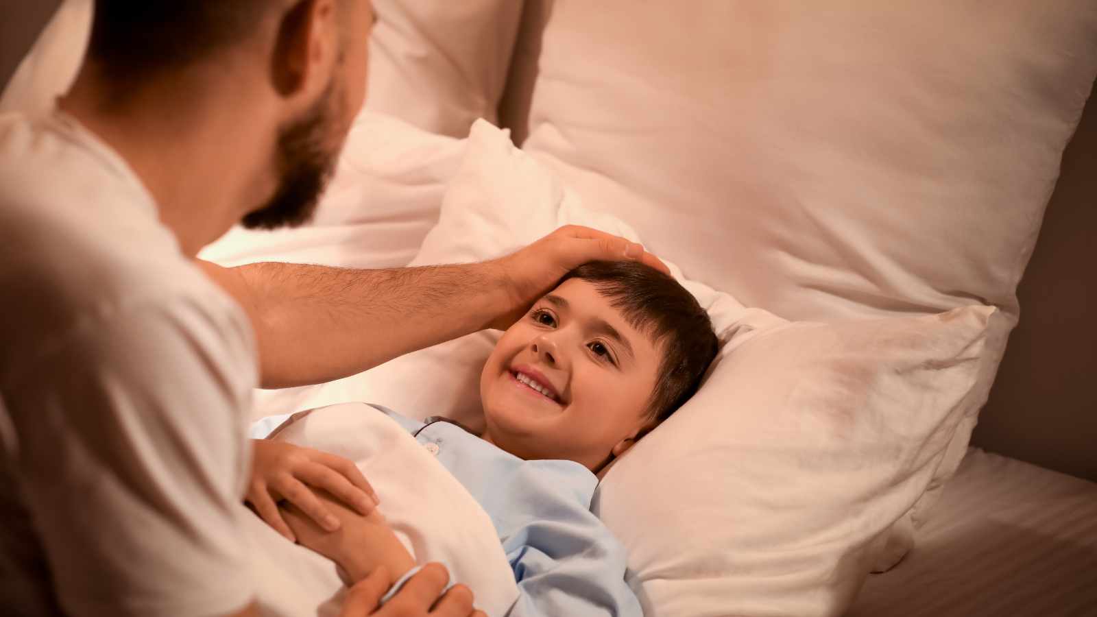A man gently pats a smiling child's head as the child lies in bed, covered with a white blanket and resting against white pillows.