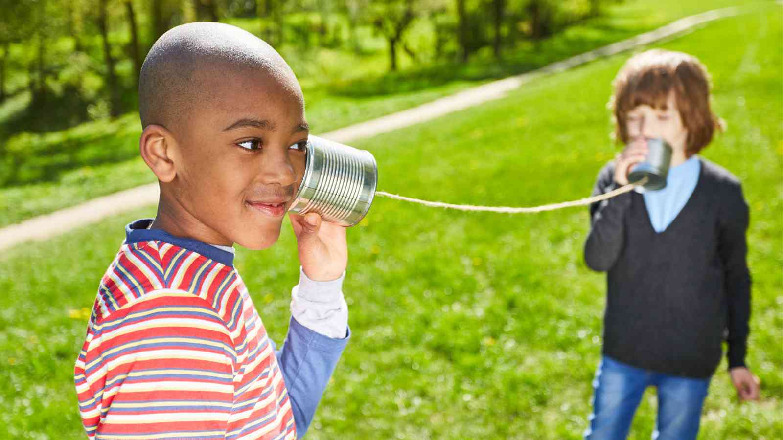 Two children in a grassy field communicate using a homemade tin can and string phone.