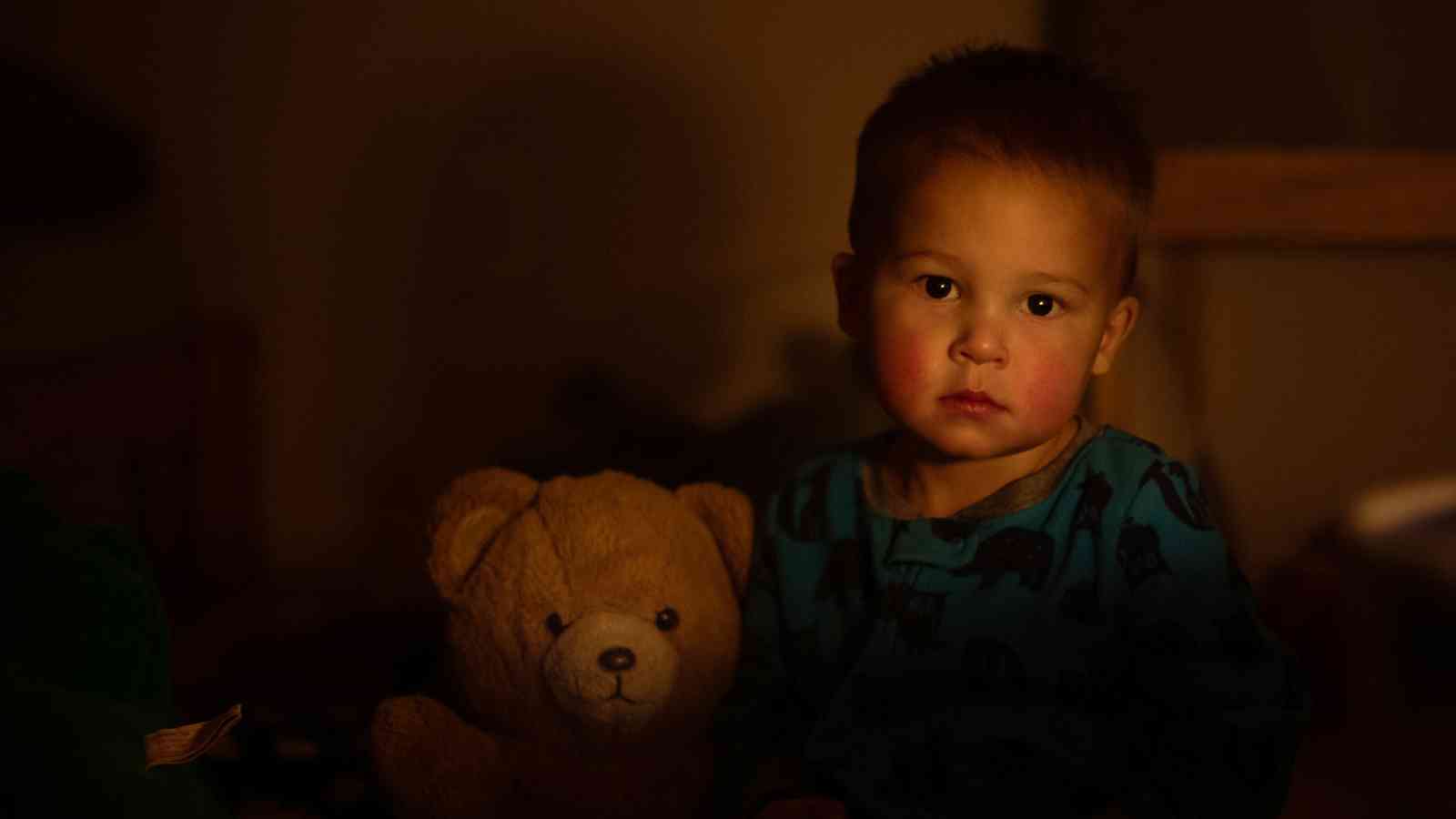 A young child sits in dim lighting next to a teddy bear, wearing a blue shirt with a pattern.