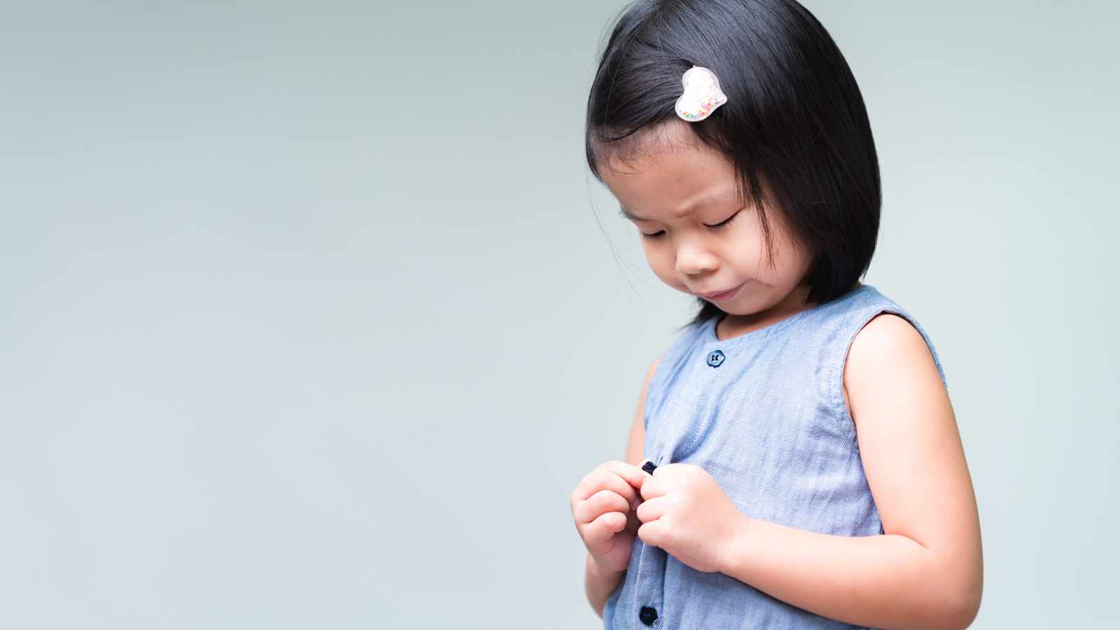 Young girl with dark hair and a hairclip buttons her blue dress, looking down with a neutral expression against a light background.
