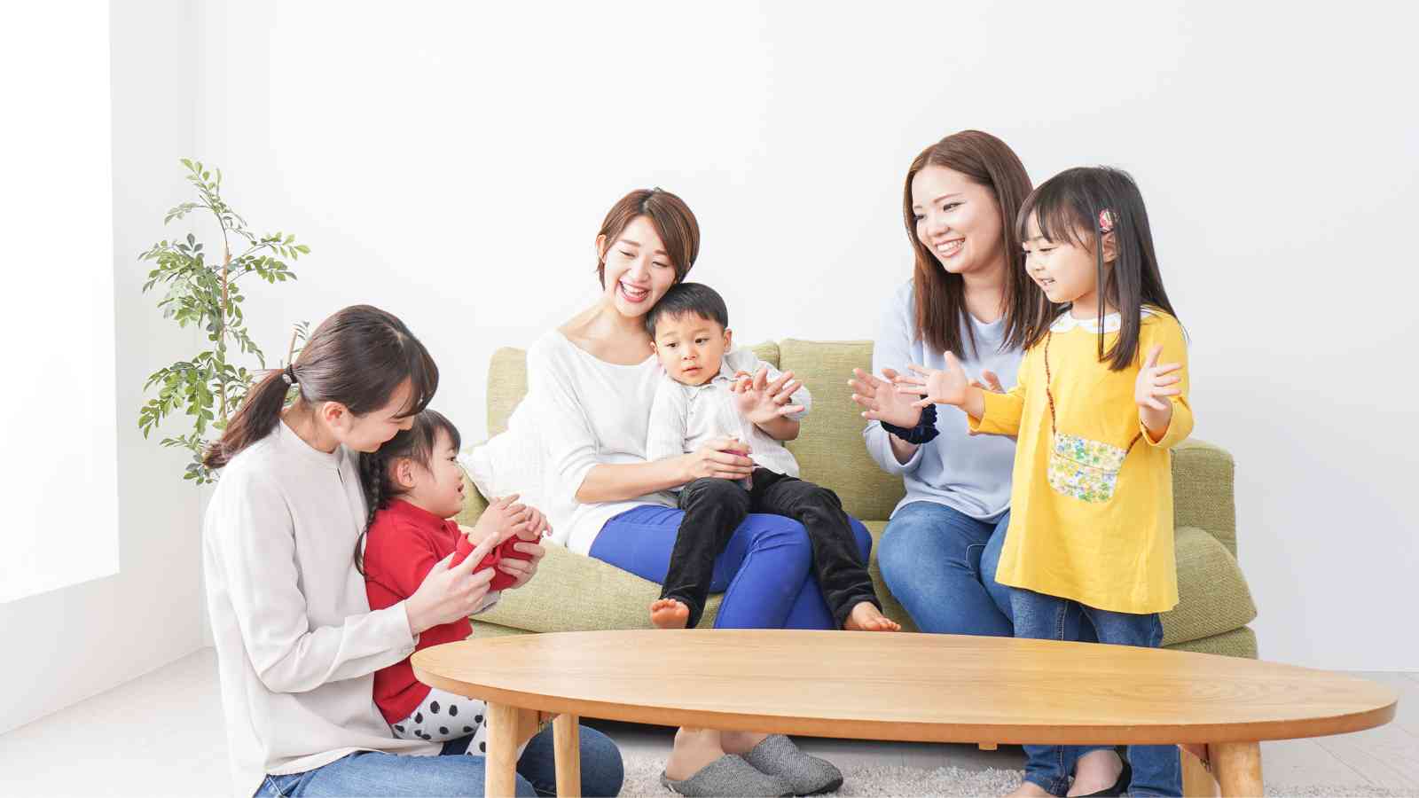 Three women and three children sit and interact happily around a wooden coffee table in a bright room with a small plant.