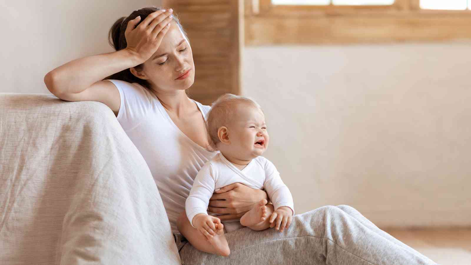 A tired woman sits on a sofa holding a crying baby, with her hand on her forehead.