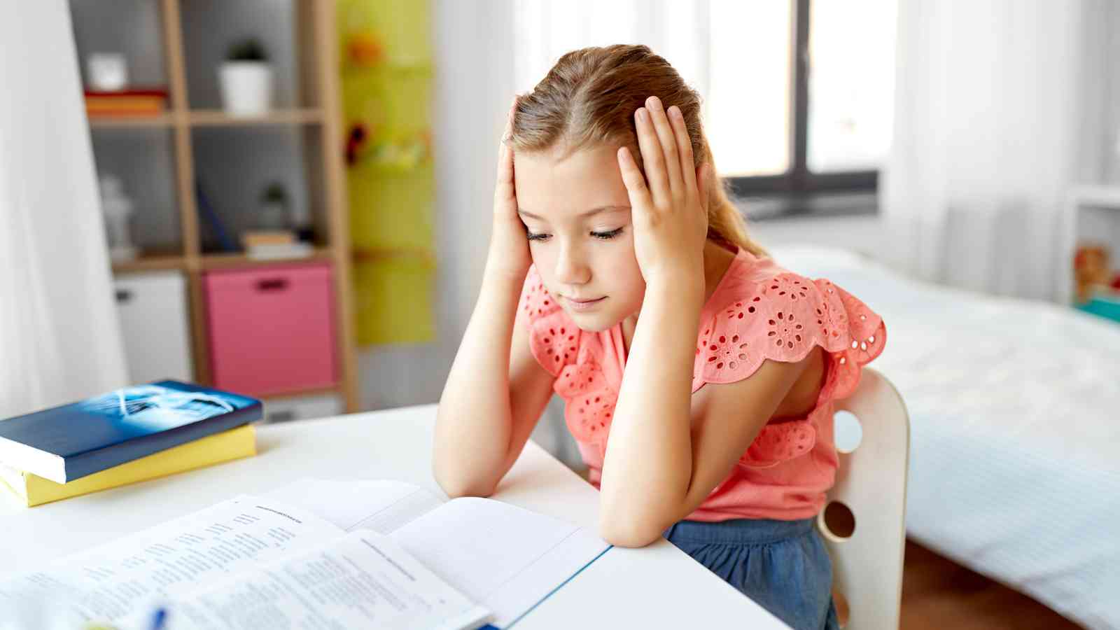 A girl sits at a table with her hands on her head, looking at an open book. Shelves and a window are in the background.