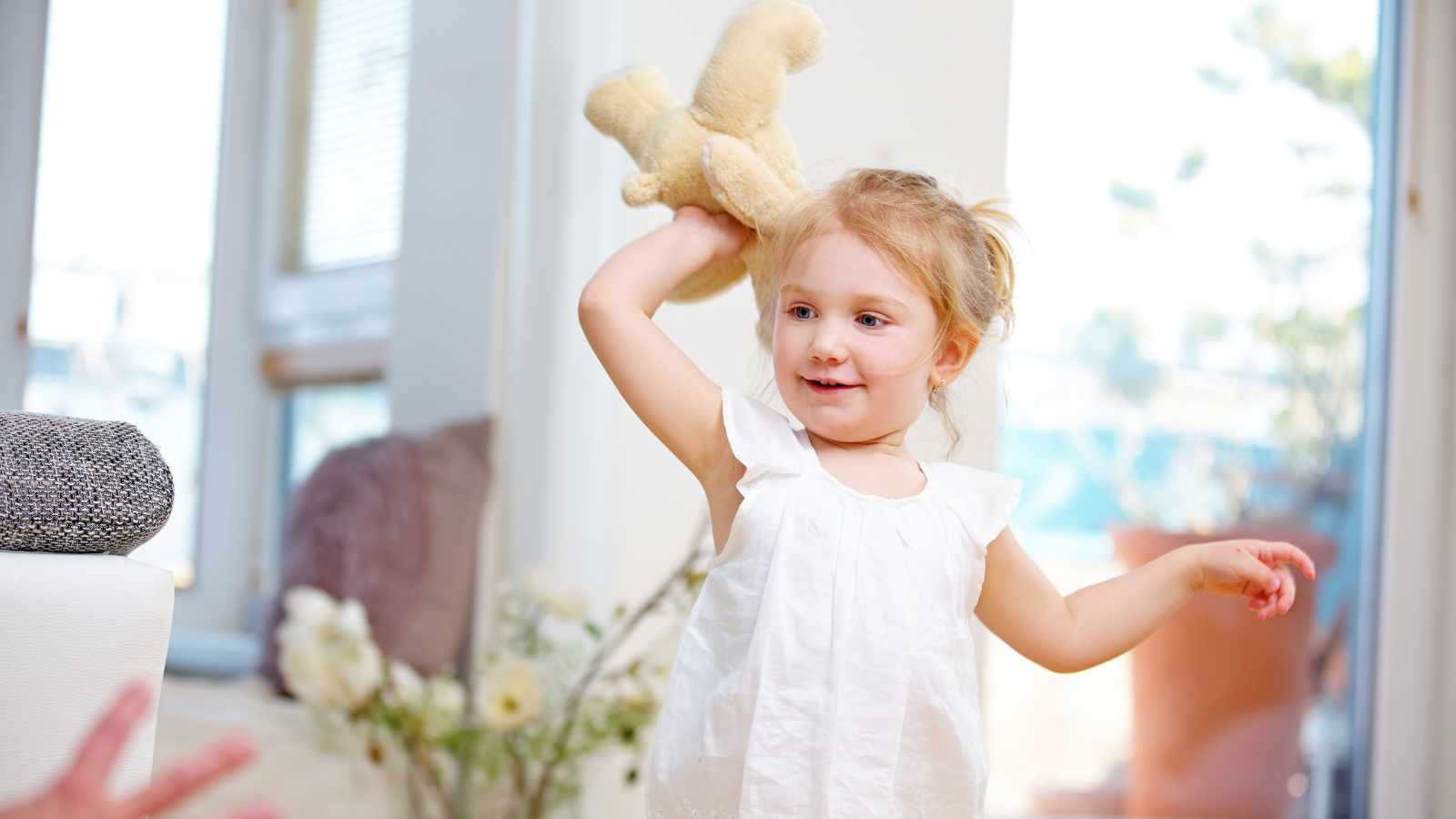 A young girl in a white dress is playing with a teddy bear indoors. She is smiling and standing near a window with flowers in the background.