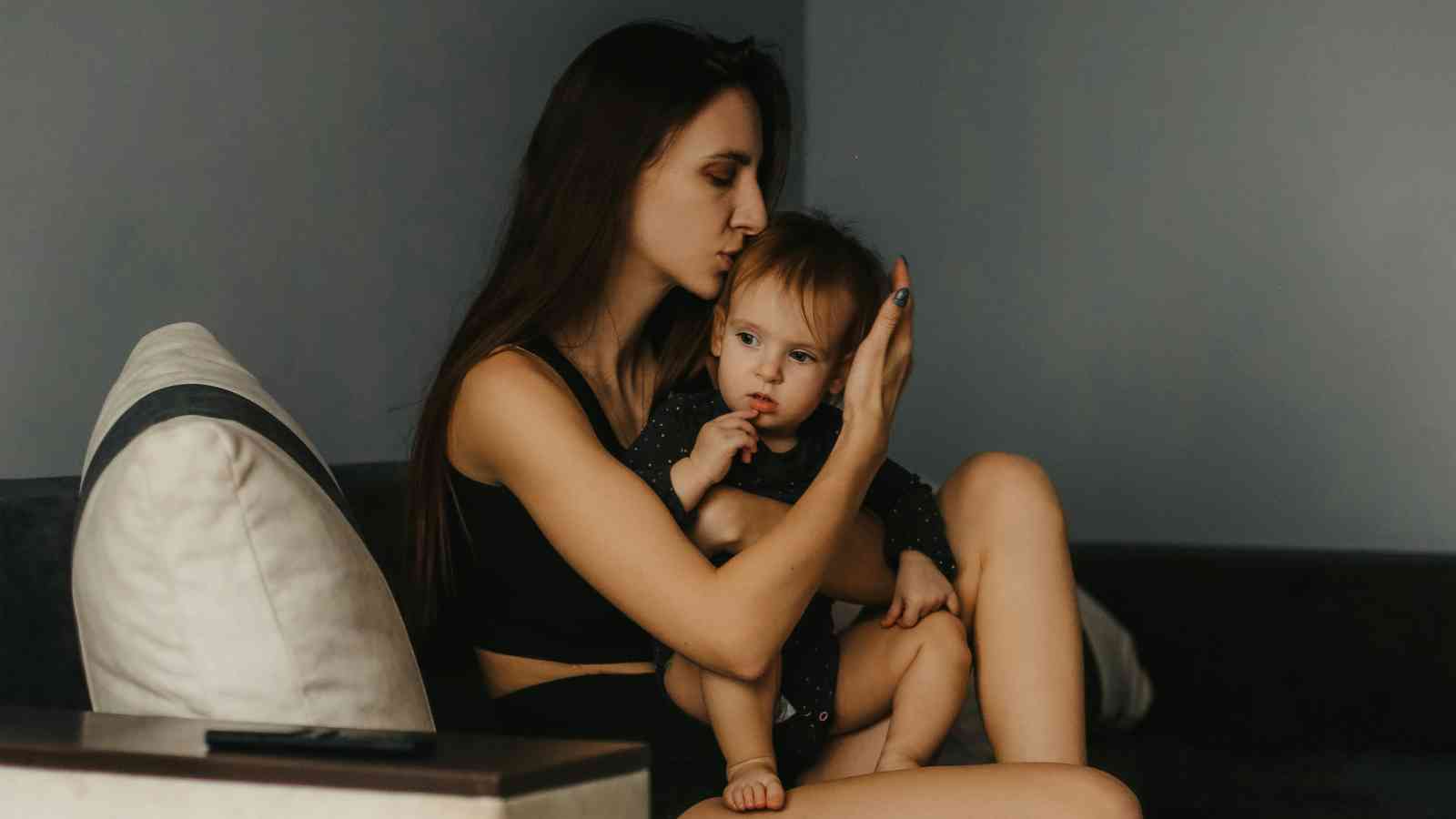A woman with long hair holds a small child on a couch, gently kissing the child's head in a dimly lit room.