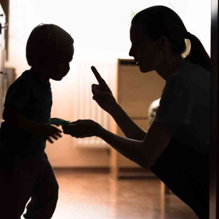 Silhouetted image of an adult kneeling, raising a finger towards a child holding something, in a hallway with backlighting from a window.