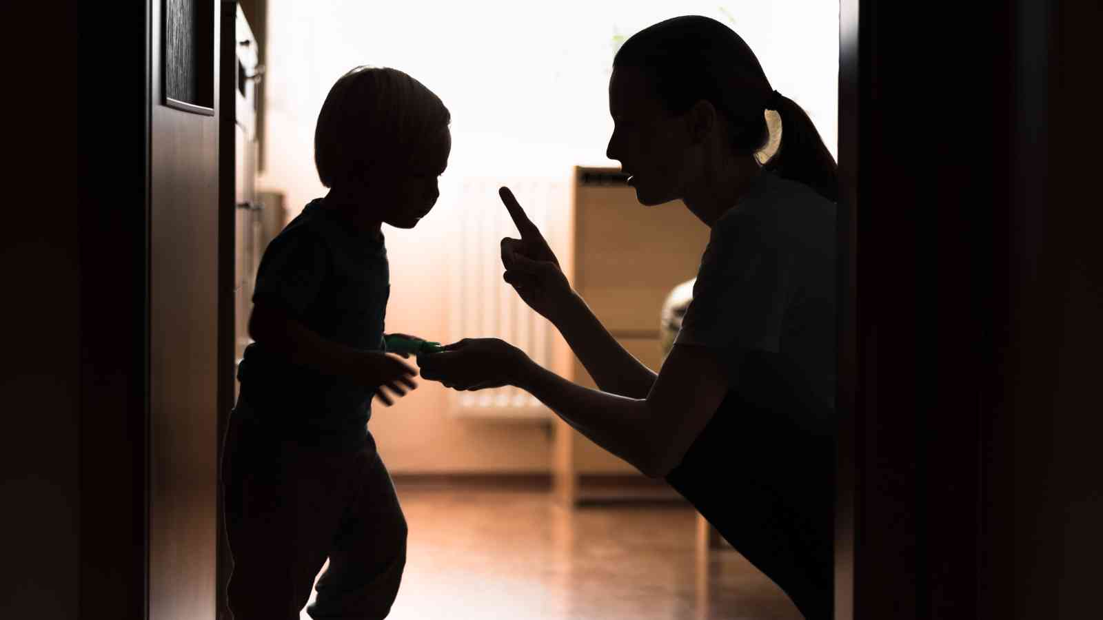 Silhouetted image of an adult kneeling, raising a finger towards a child holding something, in a hallway with backlighting from a window.