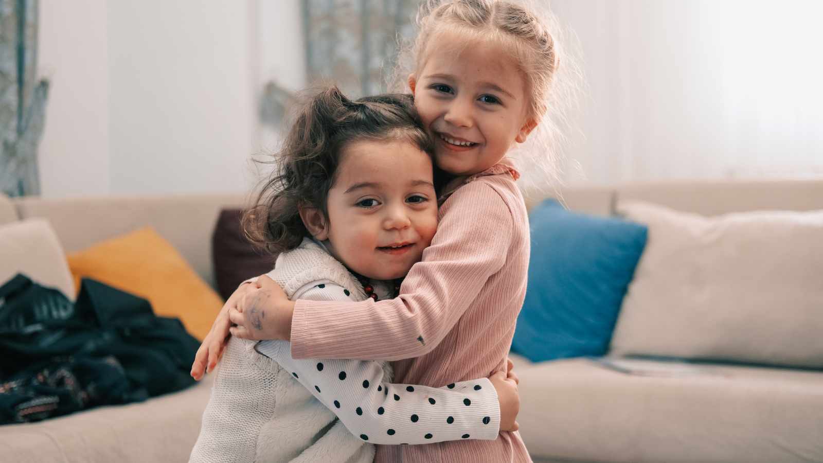 Two children smiling and hugging in a living room with a beige couch and colorful cushions in the background.