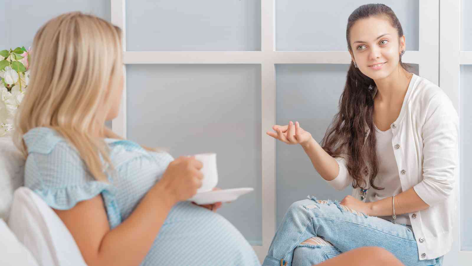 Pregnant woman holding a cup and sitting on a sofa, talking to another woman in a white cardigan and jeans.