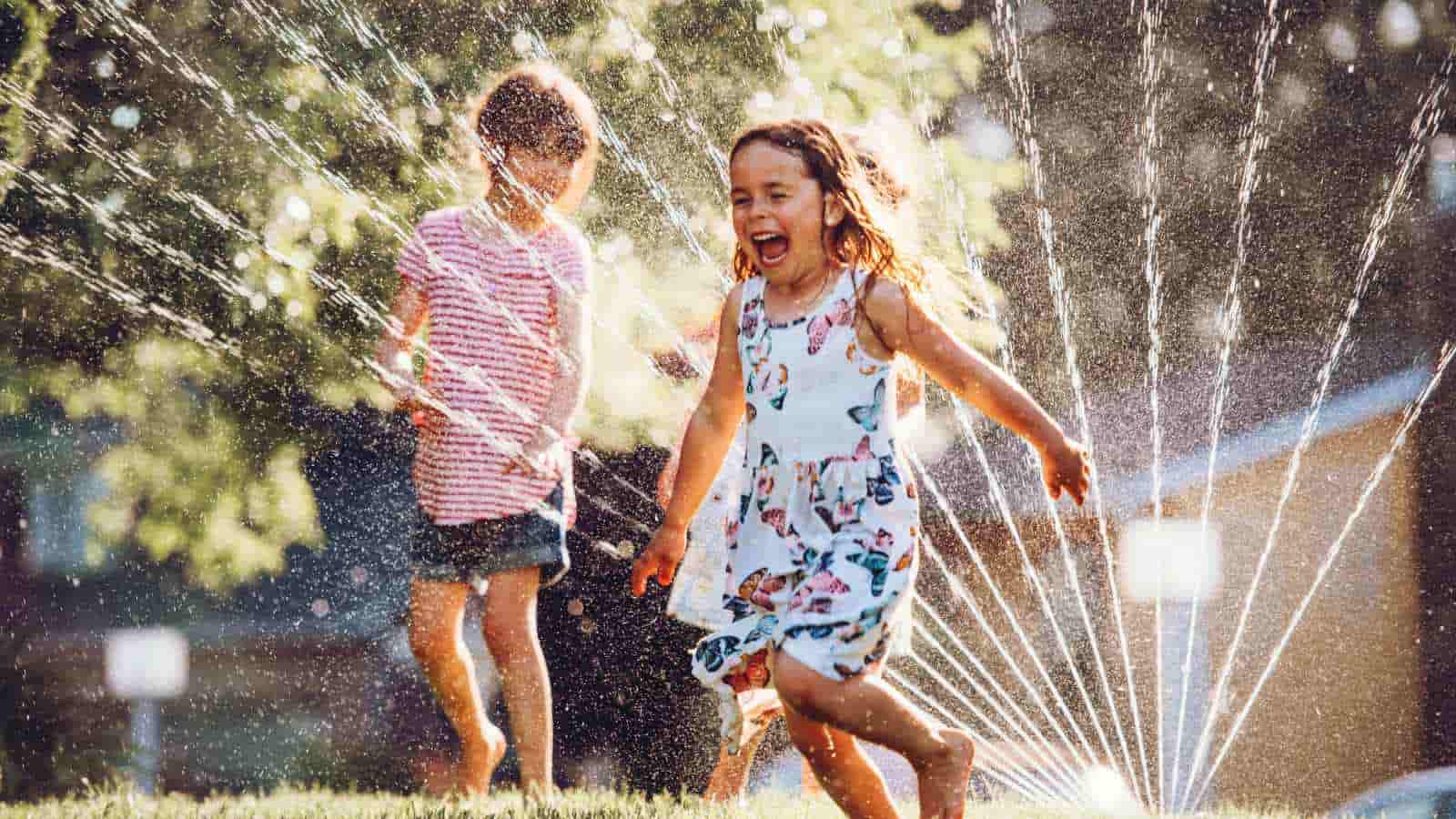 Two young children play joyfully in a garden sprinkler on a sunny day, with water droplets sparkling in the sunlight.