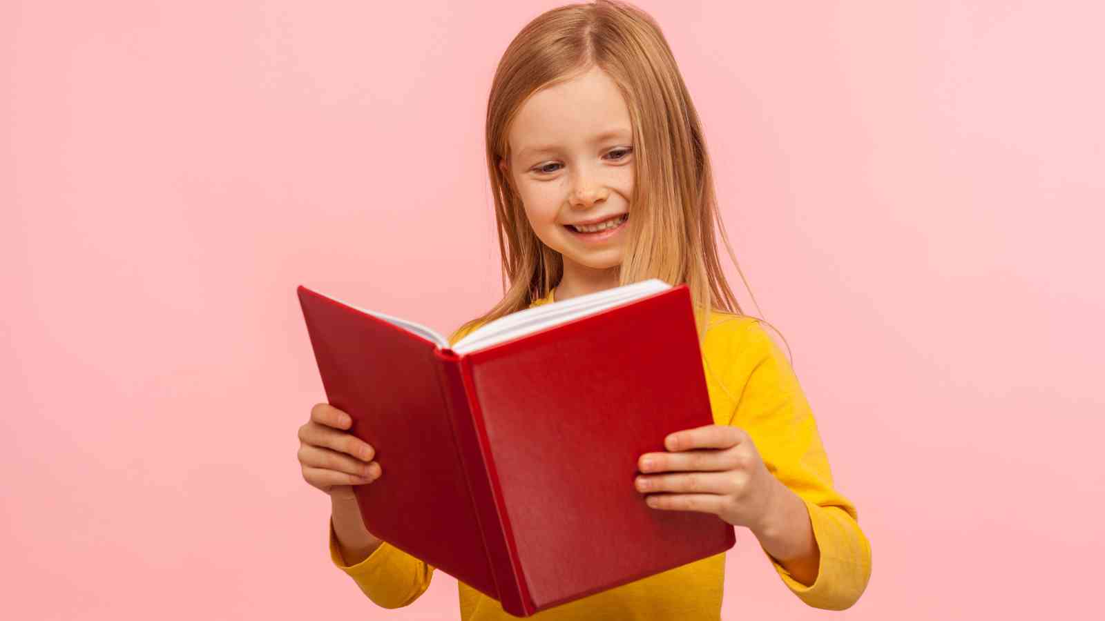 A young girl in a yellow shirt smiles while reading a red book against a pink background.