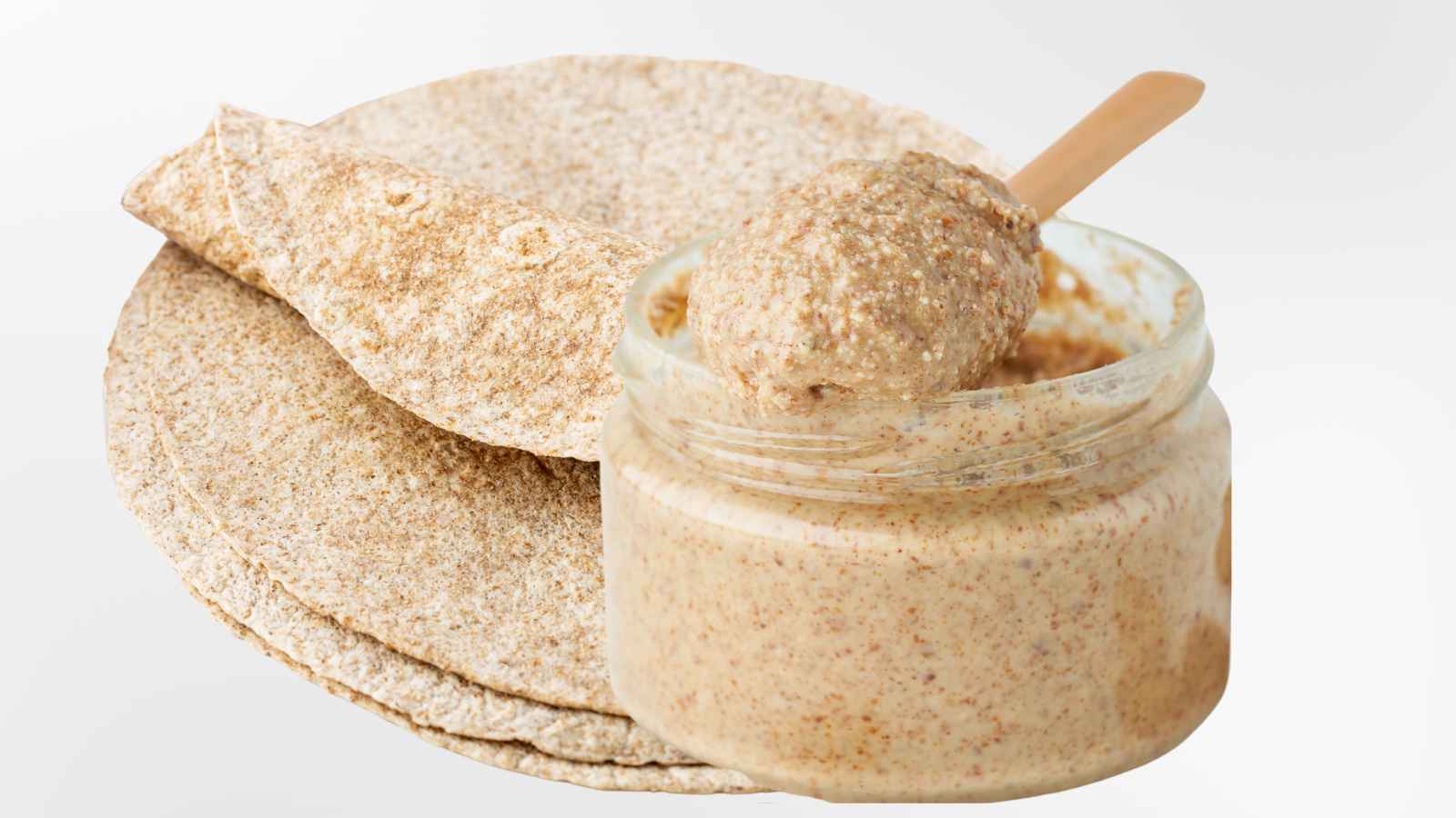 A jar of almond butter with a spoon, next to stacked flatbreads made from whole grains.