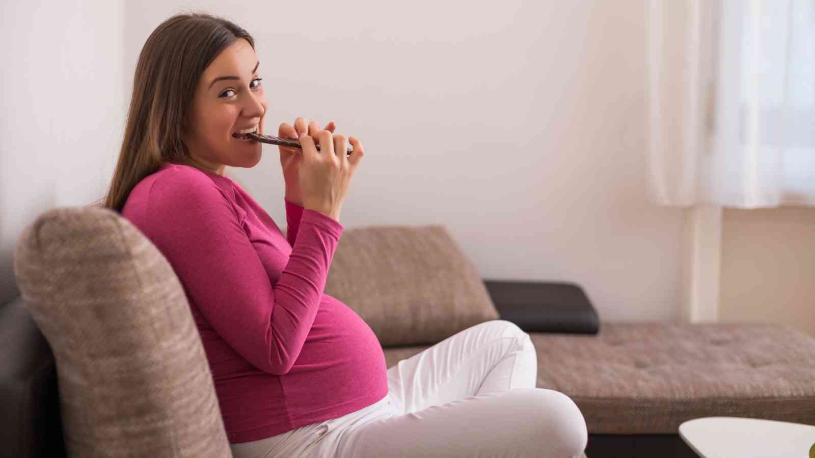 Pregnant woman in a pink top and white pants sits on a sofa, smiling and holding a chocolate bar.
