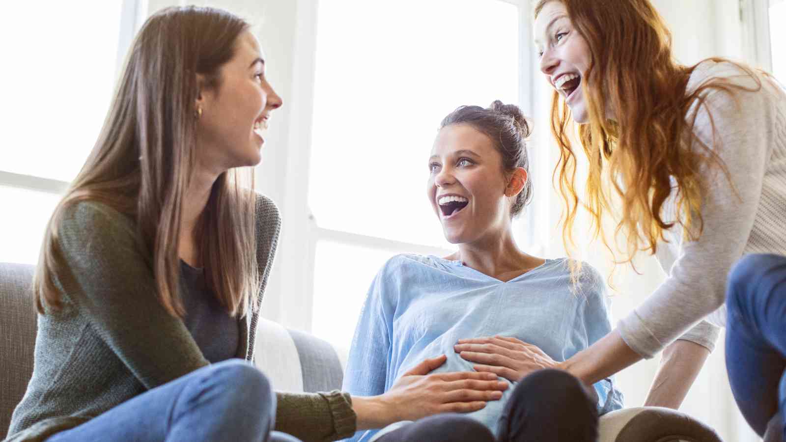 Three women sitting on a couch, one pregnant, smiling and touching her belly.