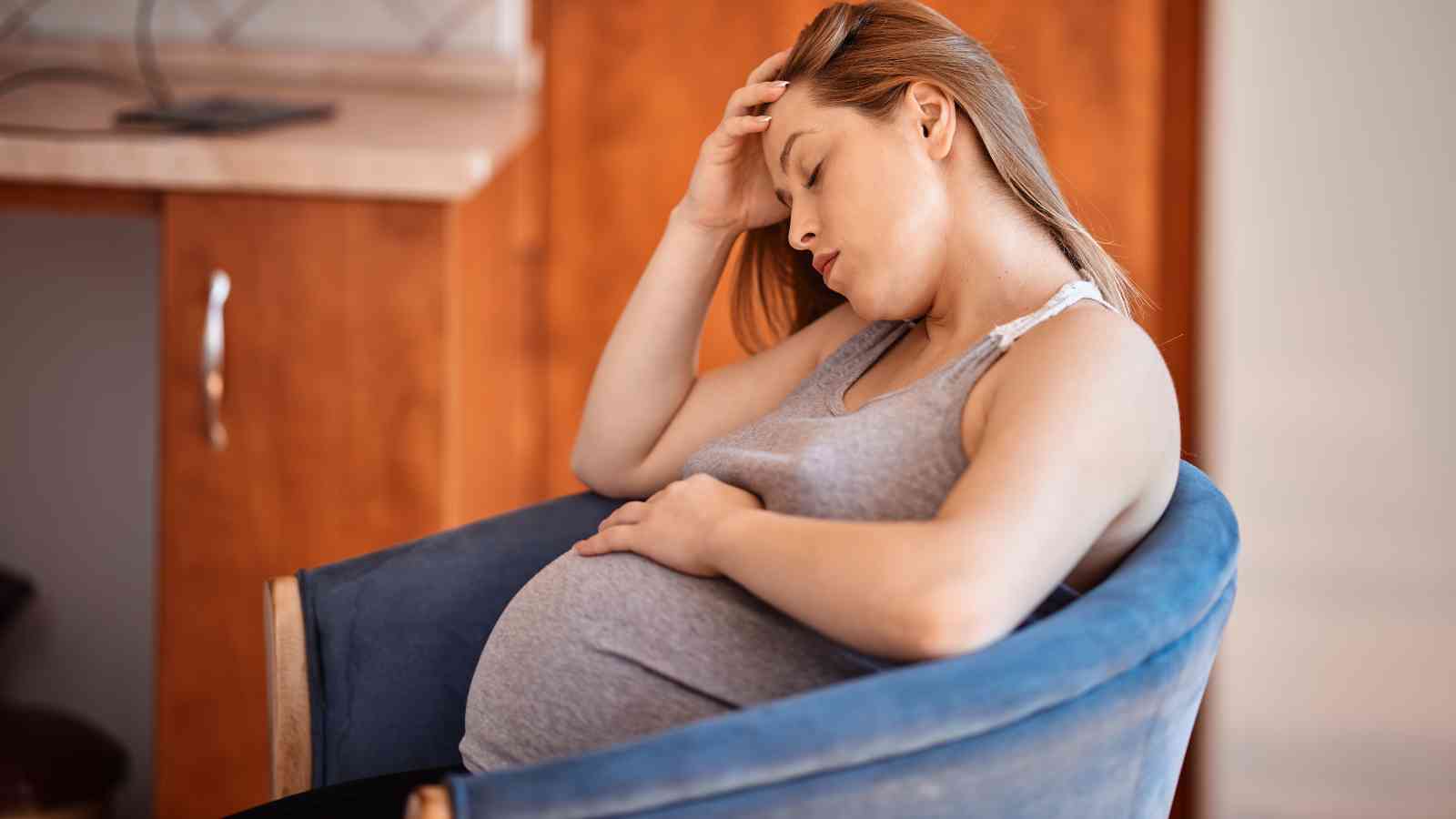 Pregnant woman in a gray tank top sitting in a blue chair, leaning back with eyes closed and hand on forehead.