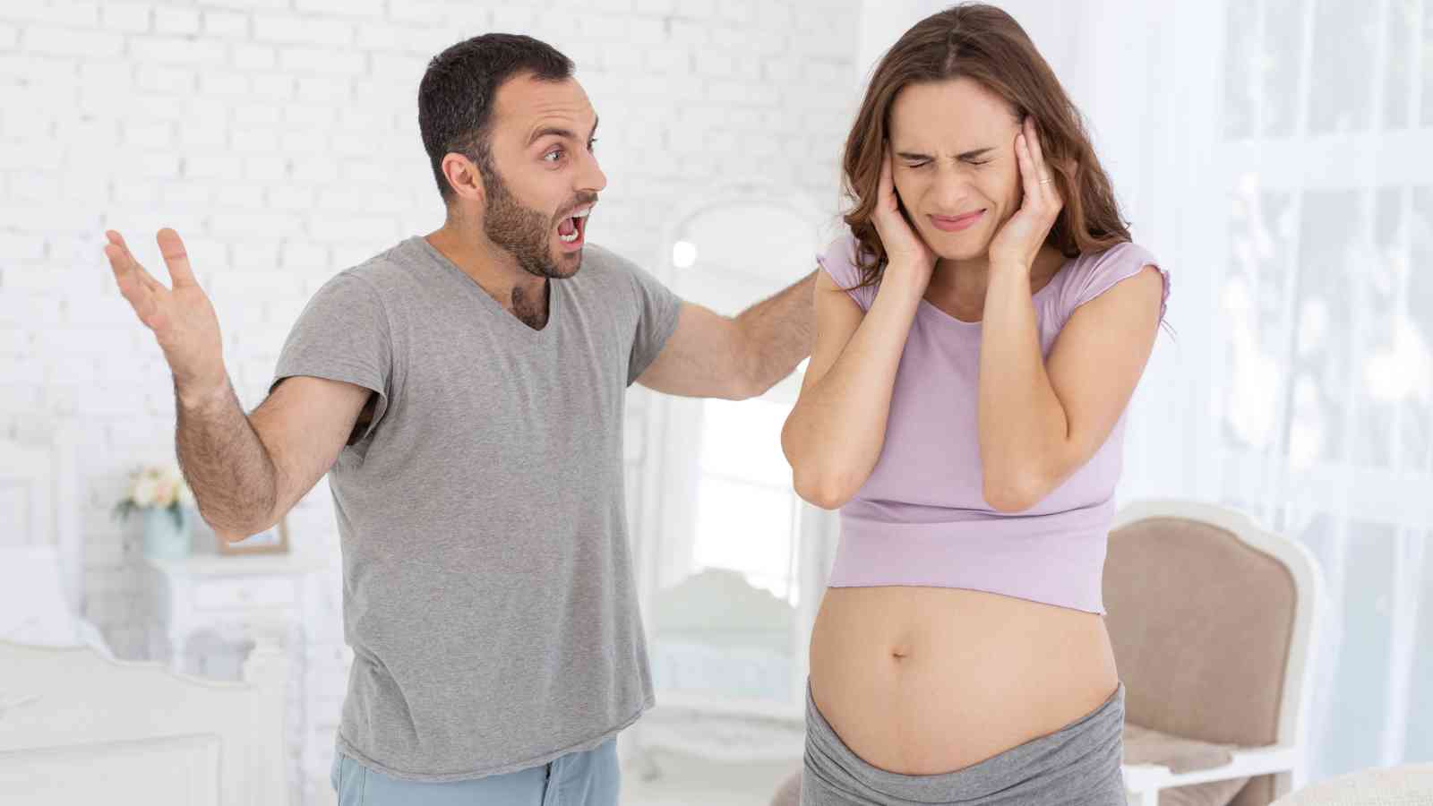 A man is yelling with raised arms, while a pregnant woman covers her ears and turns away, both standing in a bright room.