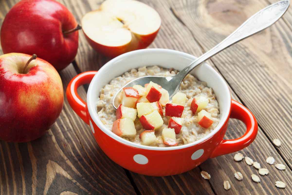 A bowl of oatmeal topped with diced apples, with a spoon inside. Two whole apples and a sliced apple half sit on a wooden table next to scattered oats. The bowl is red with white dots.