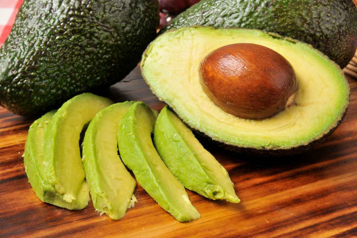 Whole and halved avocados with several slices on a wooden surface. The seed is visible in the halved avocado.