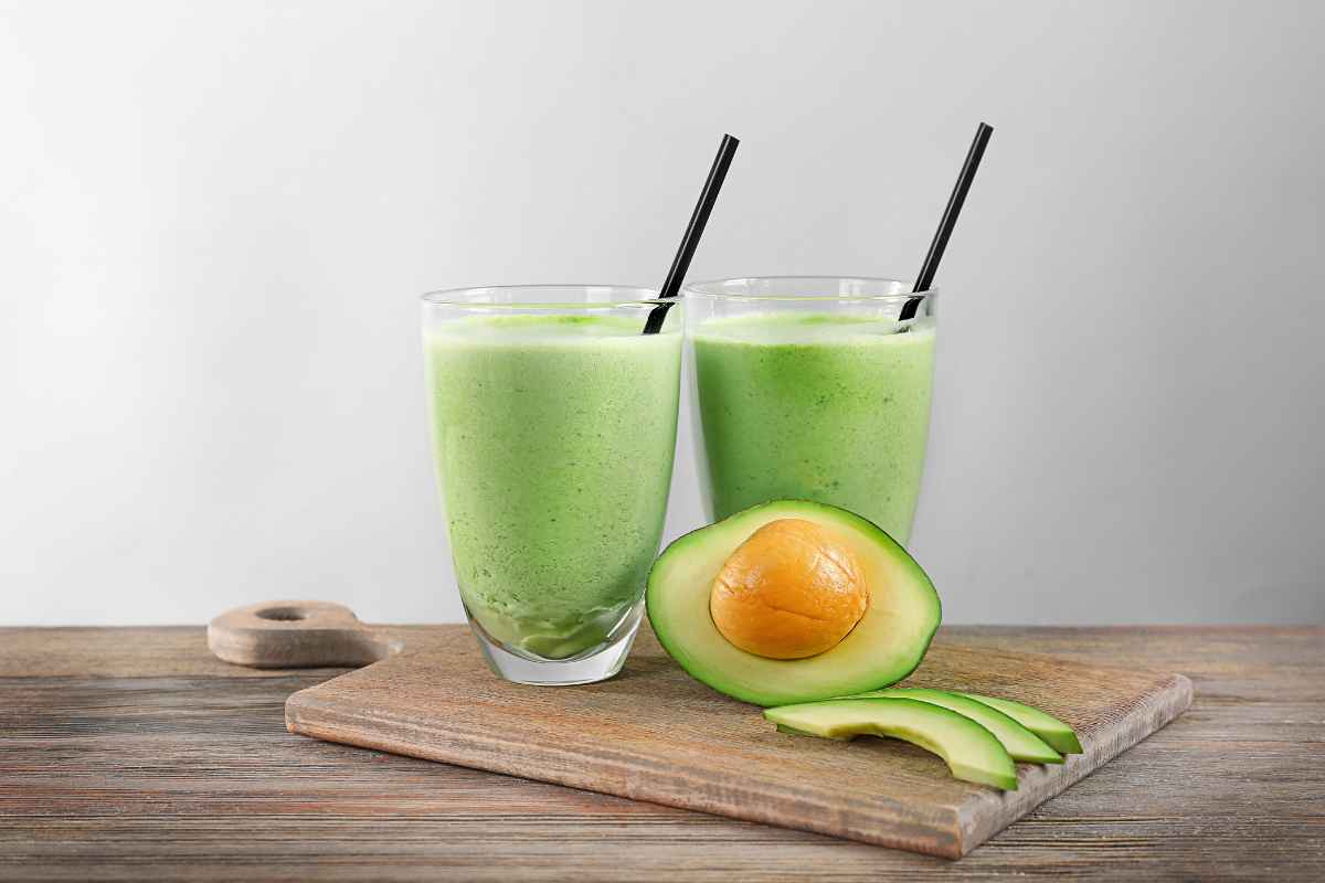 Two glasses of avocado smoothie with black straws on a wooden board, next to a halved avocado with sliced pieces.