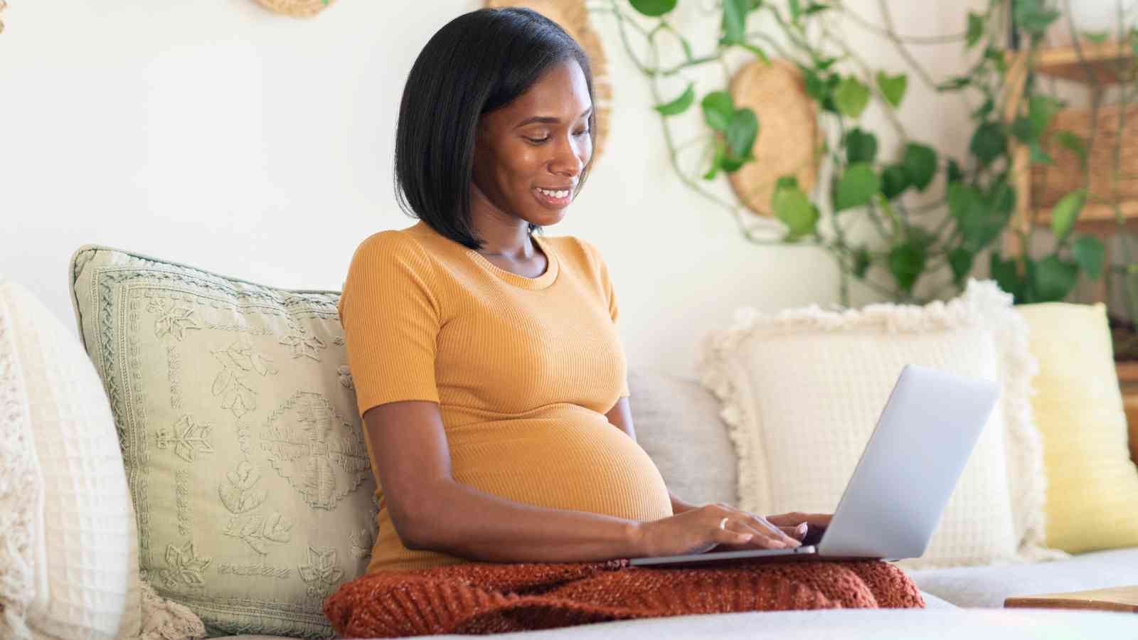 Pregnant woman sitting on a couch, using a laptop. She is smiling and surrounded by pillows and plants.