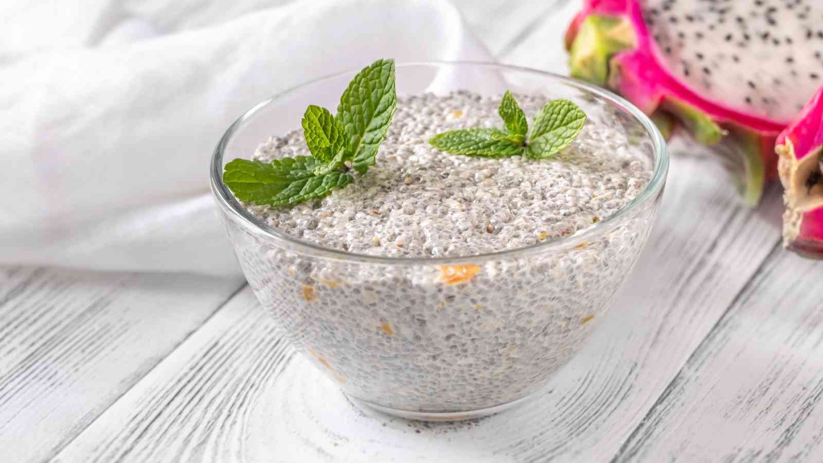 A glass bowl of chia pudding garnished with mint leaves, placed on a white wooden surface with a dragon fruit half visible in the background.