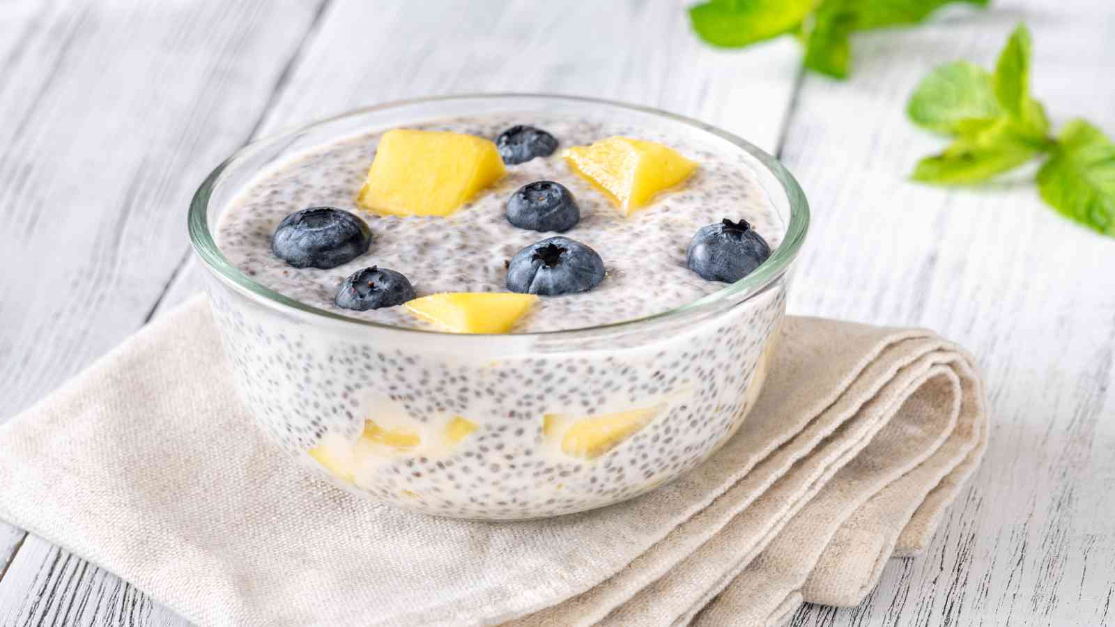 A bowl of chia pudding topped with blueberries and pineapple chunks, placed on a folded beige cloth on a light wooden surface, with green leaves in the background.