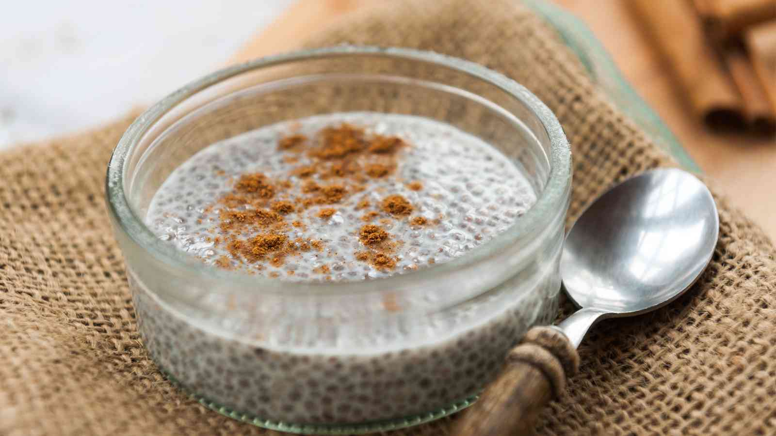 Glass bowl of chia seed pudding topped with cinnamon, placed on a burlap surface, next to a metal spoon with a wooden handle.