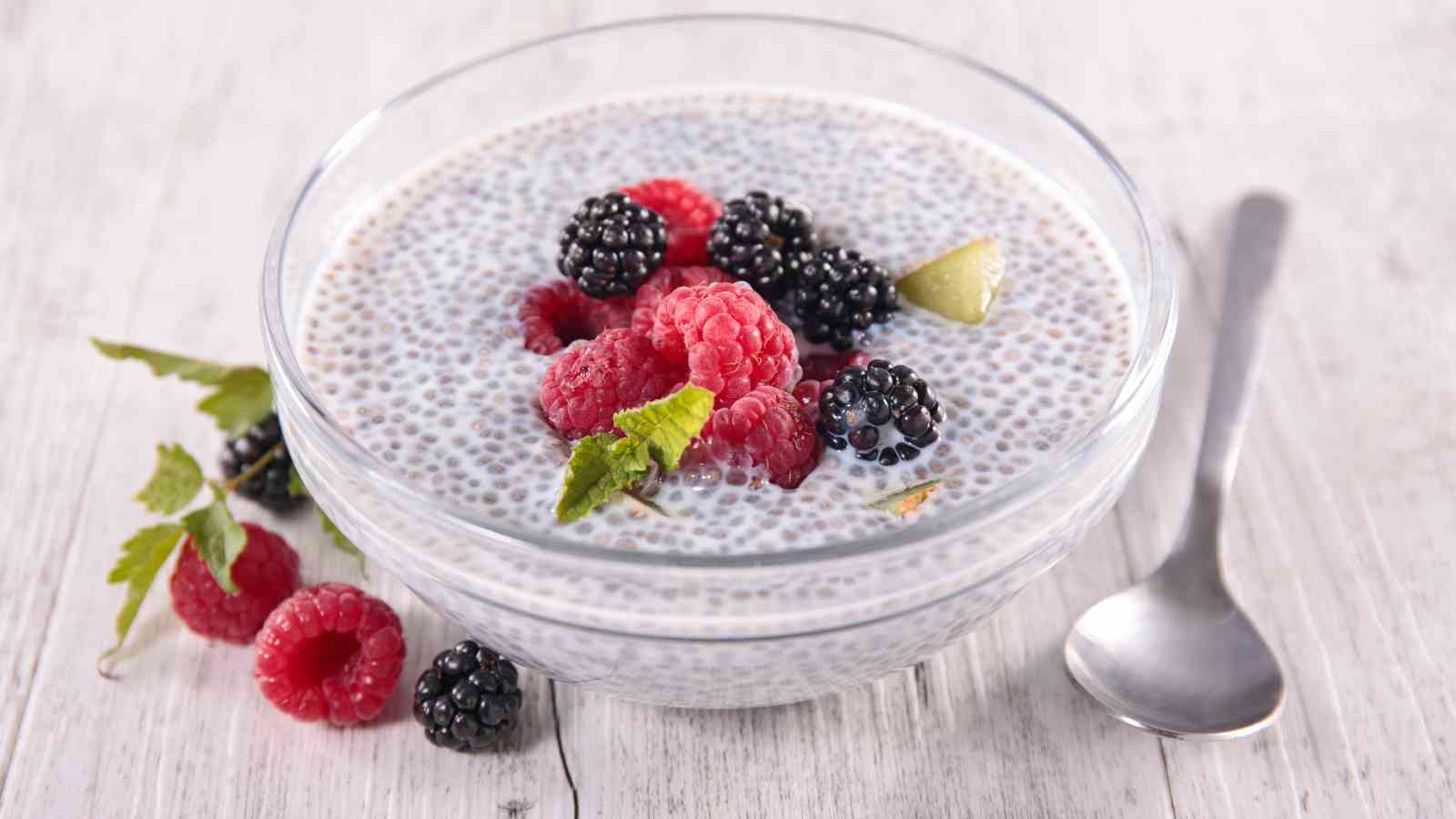 A glass bowl of chia pudding topped with raspberries, blackberries, and a mint leaf. A spoon is placed beside the bowl on a light wooden surface.