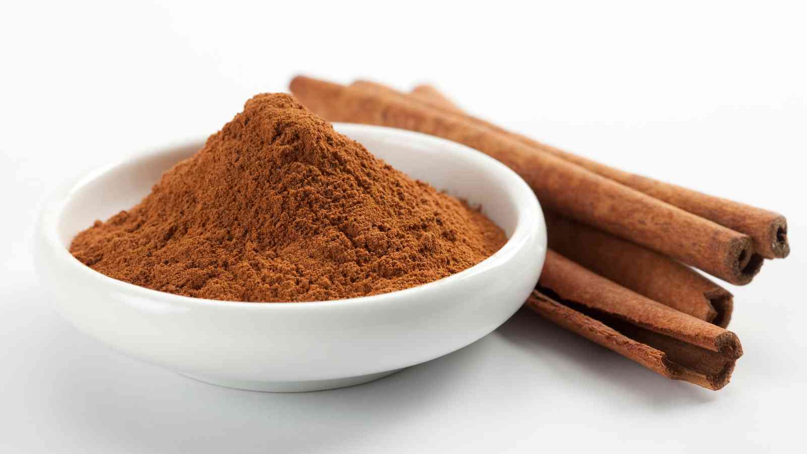 A white bowl filled with ground cinnamon, placed next to three cinnamon sticks on a white background.