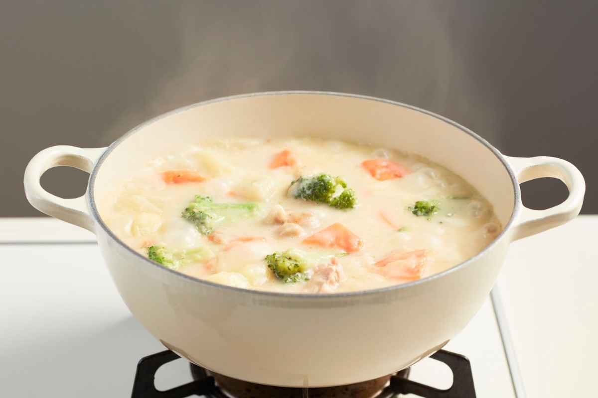 A pot of creamy soup with visible pieces of broccoli, carrots, and cauliflower, simmering on a stove with steam rising.