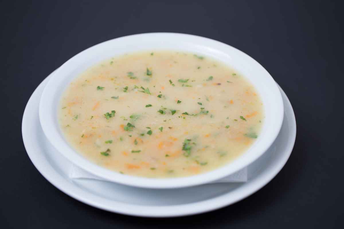 A bowl of creamy chicken soup with small pieces of herbs and vegetables, served on a white plate against a dark background.