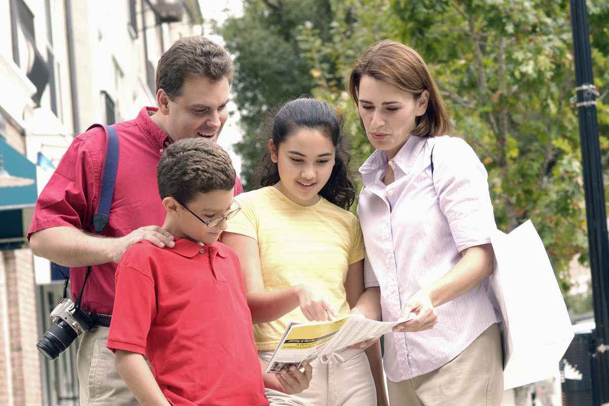 A family outdoors, two adults and two children, look at a map together. One adult holds a camera. Trees and buildings are visible in the background.
