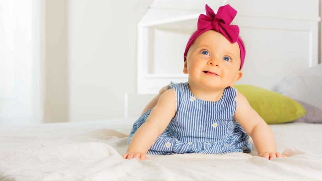 A baby wears a pink bow headband and a blue striped dress as she lies on the bed, gazing upward.