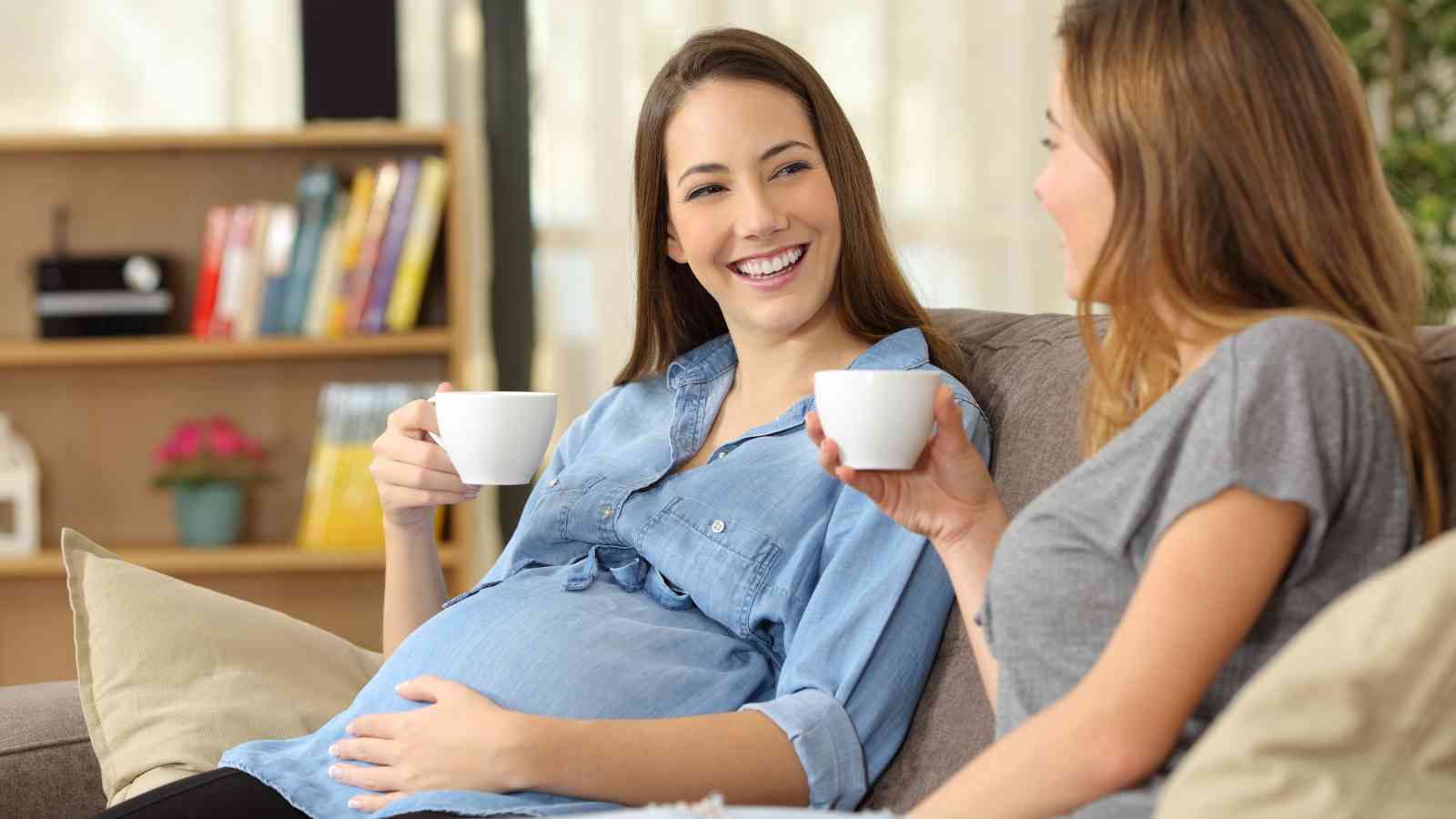Two women sitting on a couch, both holding white mugs. One woman is pregnant, touching her belly, while they engage in conversation. Bookshelves are visible in the background.