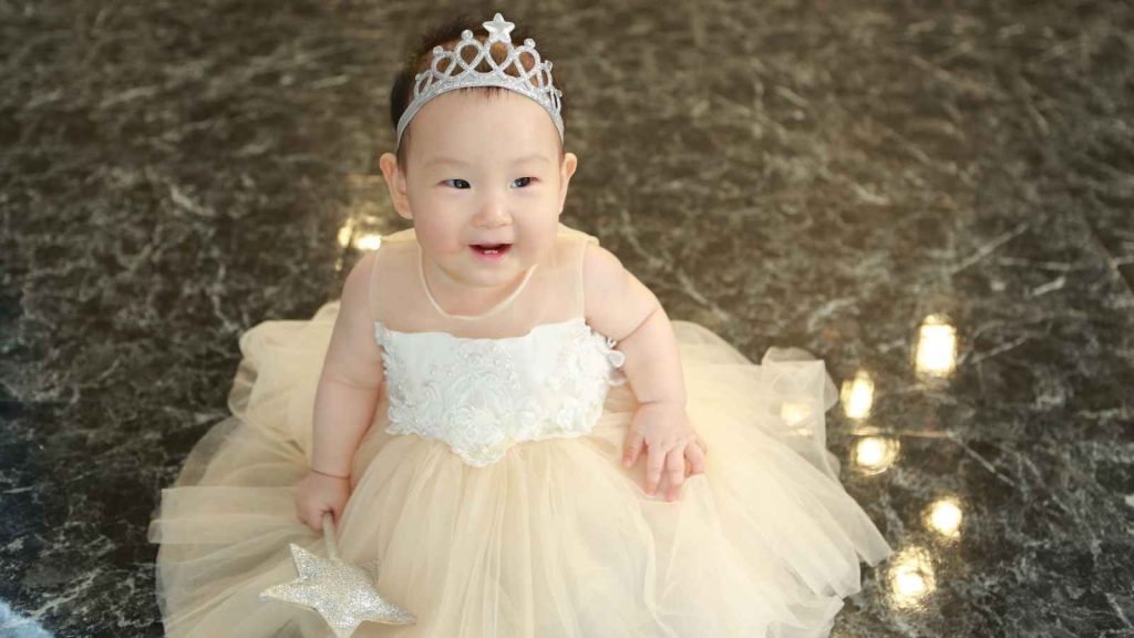 A baby wearing a white dress and silver tiara sits on a marble floor, holding a star-shaped toy.
