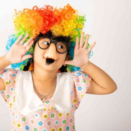 Child wearing a colorful curly wig and joke glasses with a fake nose and mustache, making a playful pose with hands near ears, set against a plain background.